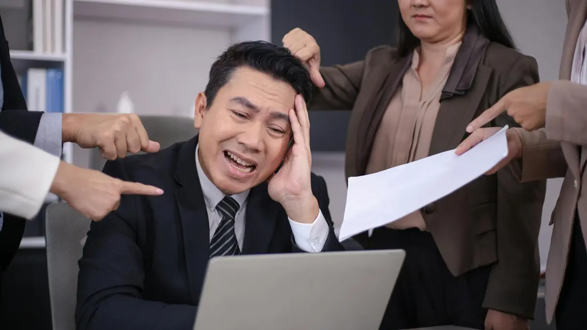 An Asian businessman looks stressed as colleagues point fingers and give him paperwork in a modern o