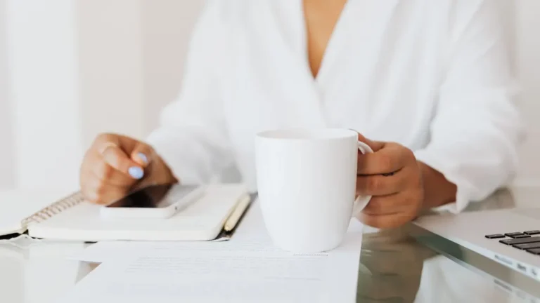 Woman holding a white mug at a desk with documents and laptop in an office.
