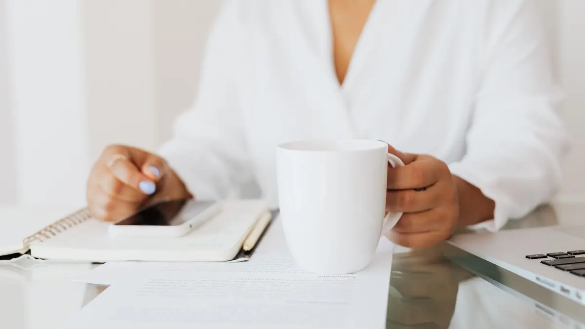 Woman holding a white mug at a desk with documents and laptop in an office.