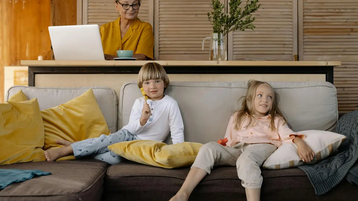 Elderly woman working on a laptop at home while grandchildren relax on the couch.